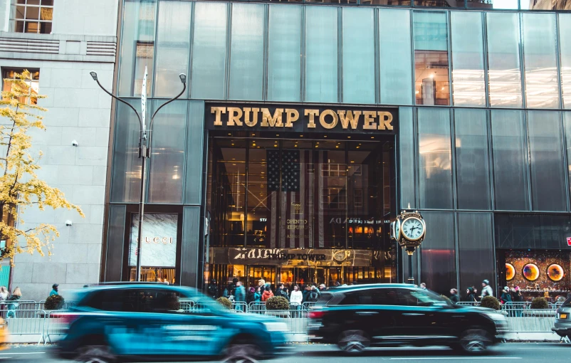 a group of cars driving down a street in front of a trump tower, by Drew Tucker, pexels contest winner, 💋 💄 👠 👗, storefront, building facing, donald trump