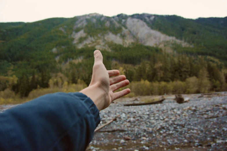 a person holding their hand out in front of a mountain, by Jessie Algie, unsplash, visual art, river in front of him, bump in form of hand, pacific northwest, raising an arm