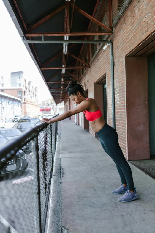 a woman standing on a sidewalk next to a fence, by Jessie Algie, unsplash, happening, sports bra, on rooftop, stretch, leaning on door