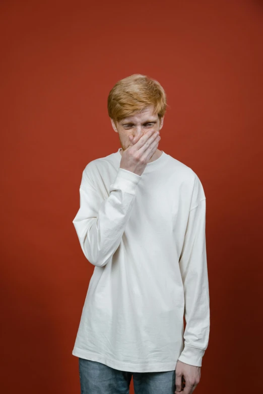 a young man standing in front of a red wall, an album cover, by Lasar Segall, trending on pexels, realism, white sweater, hr ginger, facepalm, coughing