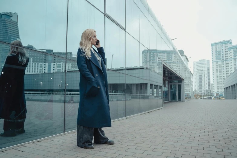 a woman standing in front of a building talking on a cell phone, a photo, by Emma Andijewska, pexels contest winner, modernism, blue skintight closed longcoat, blonde woman, paler. millions of glass-walled, moscow