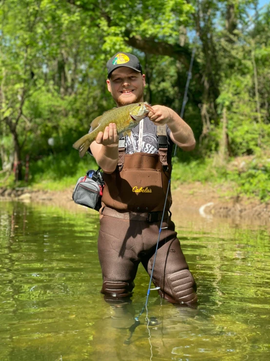 a man standing in a river holding a fish, murloc tinyfin, tyler west, wearing a fisher 🧥, big toad