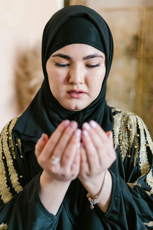 a woman in a black hijab is praying, partially cupping her hands, holding gold, contemplative, spell casting