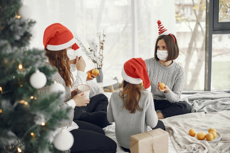 a couple of women sitting on top of a bed next to a christmas tree, by Emma Andijewska, shutterstock, surgical mask covering mouth, families playing, a group of people, red hat