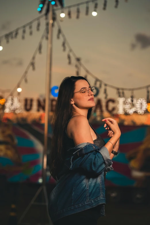 a woman standing in front of a carnival sign, by Niko Henrichon, pexels contest winner, young woman with long dark hair, girl with glasses, beautifully soft lit, panoramic view of girl