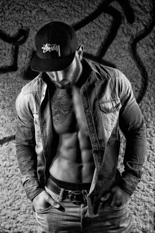 a shirtless man standing in front of a graffiti covered wall, a black and white photo, graffiti, crop shirt and strong abs, with hat, dressed in a ((ragged)), wearing a shirt and a jean