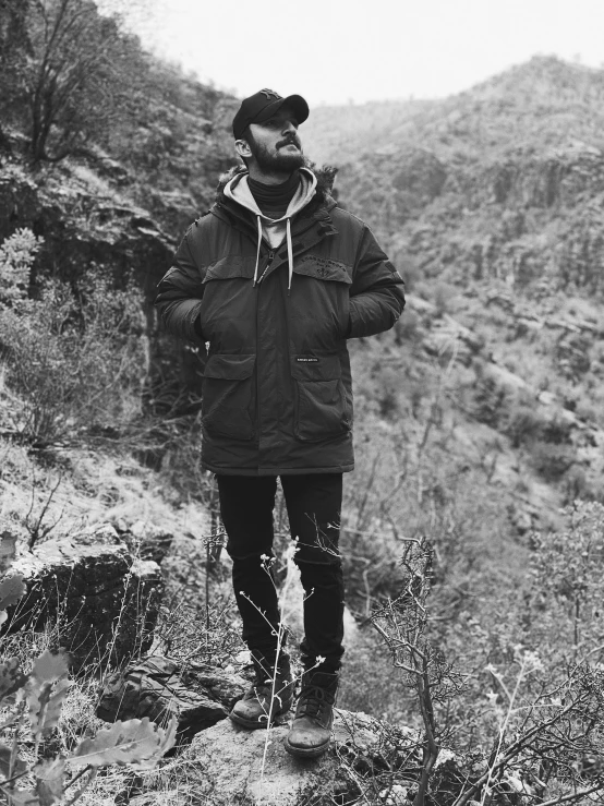 a black and white photo of a man standing on a mountain, by Kyle Lambert, wearing jacket, mac miller, amongst foliage, rugged beard