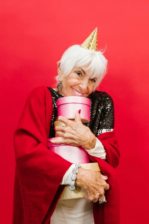 a woman in a red cape holding a pink box, by Bernie D’Andrea, pexels contest winner, a silver haired mad, wearing a party hat, hugging, holding gift