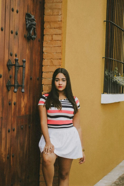 a woman standing in front of a wooden door, by Alejandro Obregón, wearing stripe shirt, white skirt, chloe bennet, full frame image