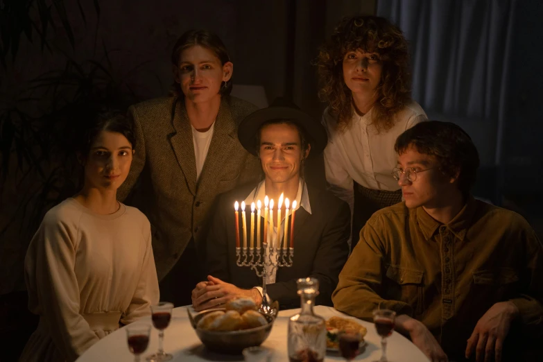 a group of people sitting around a table with candles on it, robert sheehan, hila klein, [ theatrical ]