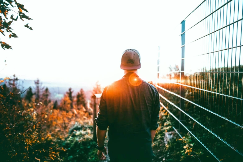 a man wearing a hat standing in front of a fence, pexels contest winner, happening, back lit, towering high up over your view, instagram picture, looking off into the distance