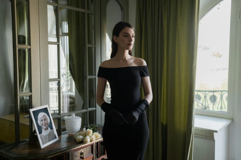 a woman in a black dress standing in front of a window, a portrait, inspired by Carlo Mense, funeral, phoebe tonkin, filmstill, ( ( theatrical ) )