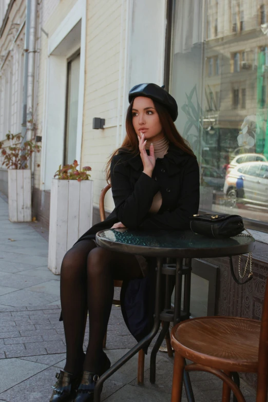 a woman sitting at a table in front of a window, lady in black coat and pantyhose, street pic, black beret, mall goth