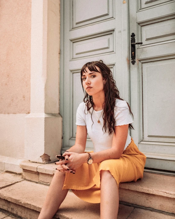 a woman sitting on the steps of a building, inspired by Valéria Dénes, trending on pexels, happening, yellow hue, brown hair and bangs, dressed in a white t-shirt, belle delphine