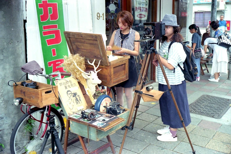 a couple of women standing in front of a camera, a silk screen, instagram, ukiyo-e, old japanese street market, tripod, movie set”, “ painting