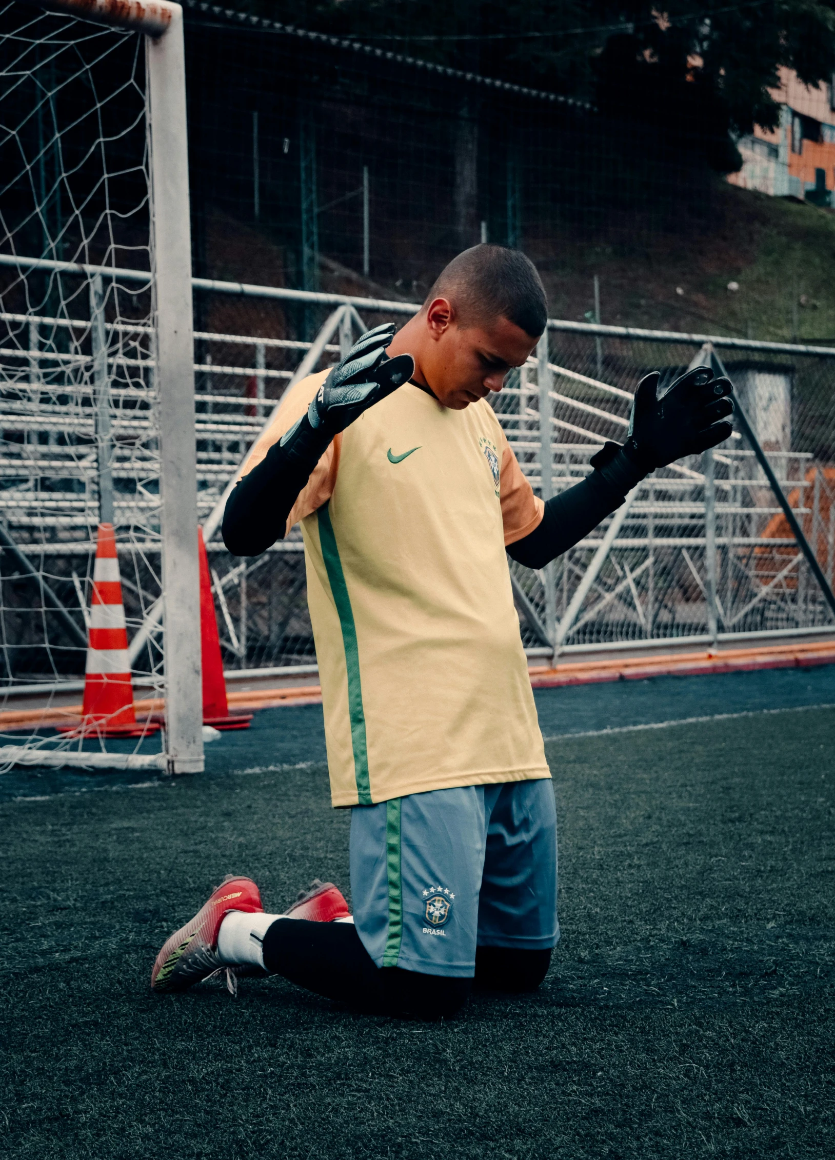 a young man kneeling on top of a soccer field, inspired by Nuno Gonçalves, pexels contest winner, square, holding his hands up to his face, low quality photo, in sao paulo