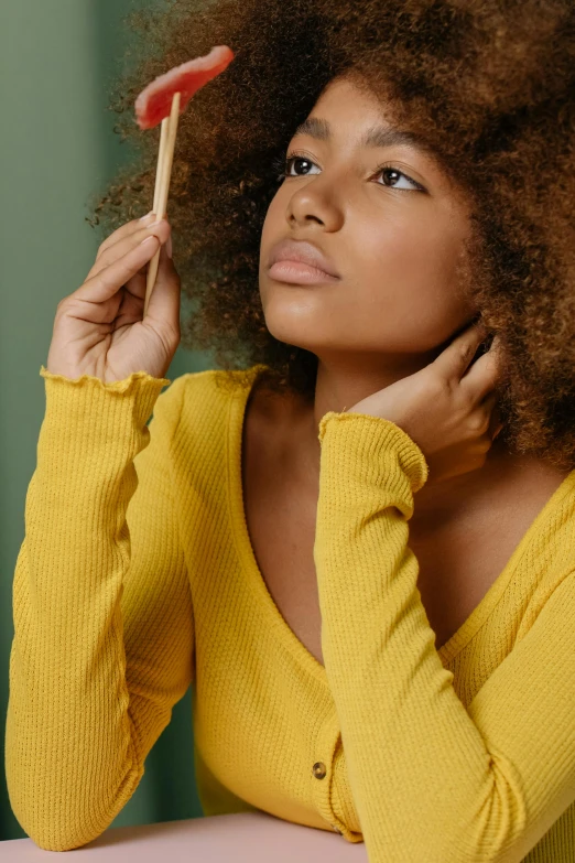 a woman sitting at a table with a lollipop lollipop lollipop lollipop lollipop lollipop lolli, trending on pexels, renaissance, wearing a yellow hoodie, natural hair, looking in mirror, thoughtful )