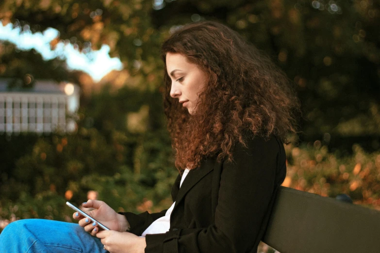 a woman sitting on a bench using a cell phone, pexels, happening, brown curly hair, using a magical tablet, avatar image, side profile shot