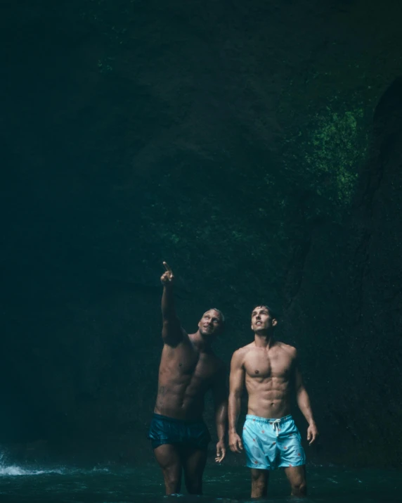 two men standing in the water in front of a waterfall, a cave painting, by Adam Dario Keel, sumatraism, homoerotic!, caves!