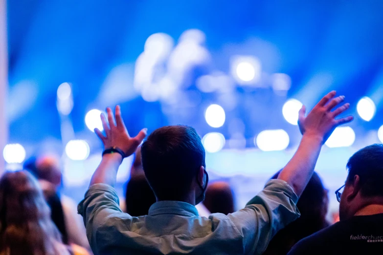 a group of people standing in front of a stage, pexels, happening, indistinct man with his hand up, listening to godly music, brightly lit blue room, a photo of a man