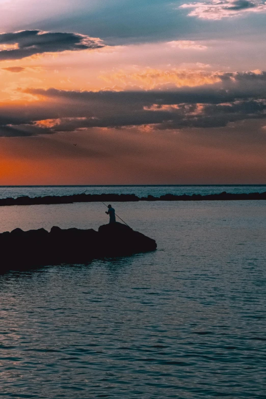 a person standing on a rock in the middle of a body of water, at sunset, fishing, in the evening