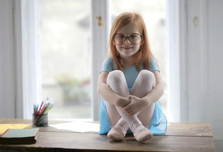 a little girl sitting on top of a wooden table, pexels contest winner, blue rimmed glasses, dressed as a ballerina, sitting in the classroom, redhead girl