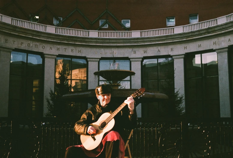 a man sitting in a chair playing a guitar, an album cover, inspired by Joseph Yoakum, unsplash, in moscow centre, in front of the house, a pilgrim, live performance