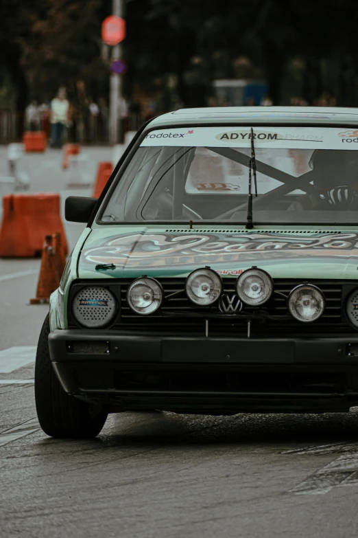 a green car driving down a street next to orange cones, pexels contest winner, photorealism, rally car, round headlights, malaysian, bmw e 3 0