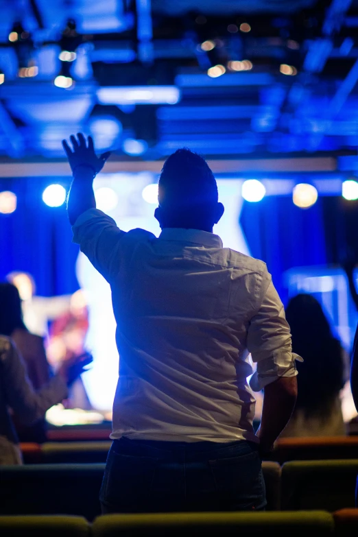 a man standing in front of a stage with his hands in the air, by Dan Content, worshipers, brightly lit blue room, on ship, pondering