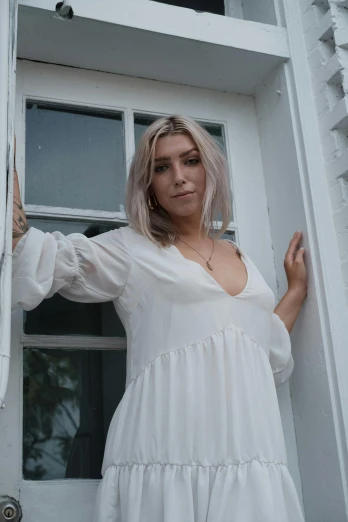 a woman in a white dress standing in front of a door, inspired by Elsa Bleda, pexels contest winner, aestheticism, young blonde woman, white backdrop, music video, looking up at camera