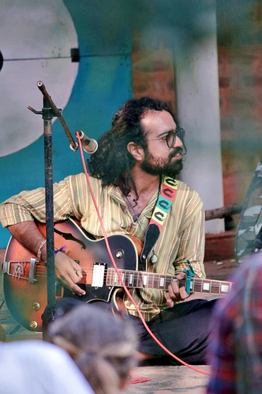 a man that is sitting down with a guitar, bengal school of art, a person at a music festival, no cropping, square, 7 0 s vibe