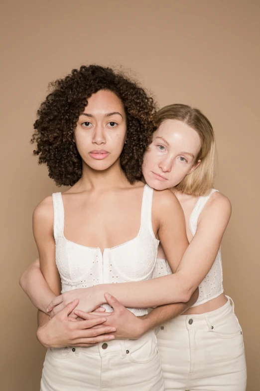 a couple of women standing next to each other, inspired by Vanessa Beecroft, trending on pexels, renaissance, mixed race woman, pale skin curly blond hair, lesbian embrace, plain background