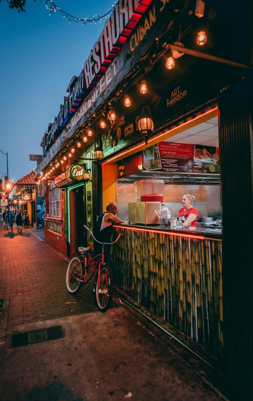 a person sitting on a bike in front of a restaurant, in the evening, getting his tacos and drink), london at night, neon shops