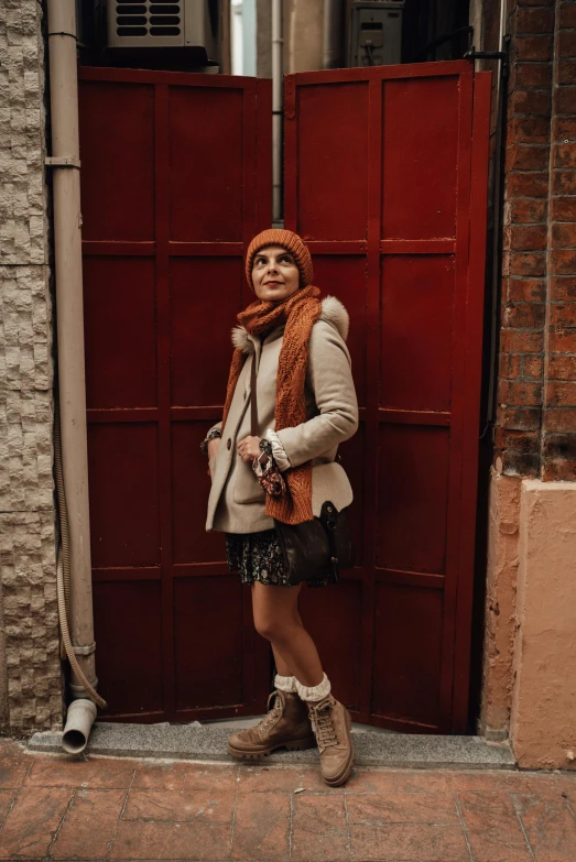 a woman standing in front of a red door, by Lucia Peka, pexels contest winner, wearing beanie, wears brown boots, orange tones, instagram post