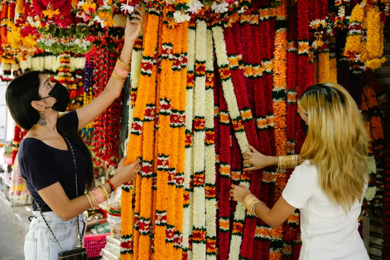 a couple of women standing in front of a wall of flowers, trending on unsplash, hurufiyya, hindu ornaments, market stalls, black and yellow and red scheme, beaded curtains