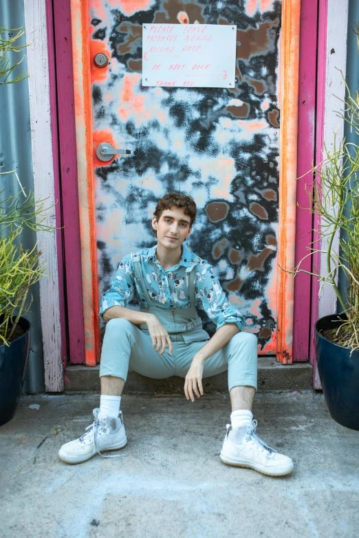 a man sitting in front of a door next to potted plants, an album cover, by Jessie Alexandra Dick, trending on pexels, graffiti, portrait of timothee chalamet, wearing a tie-dye shirt, nonbinary model, wearing overalls