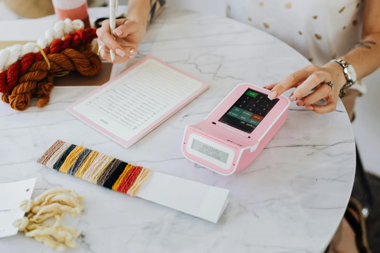 a woman sitting at a table working on a project, a cross stitch, inspired by Angelica Kauffman, pexels contest winner, cash register, pink color scheme, scanning items with smartphone, product view