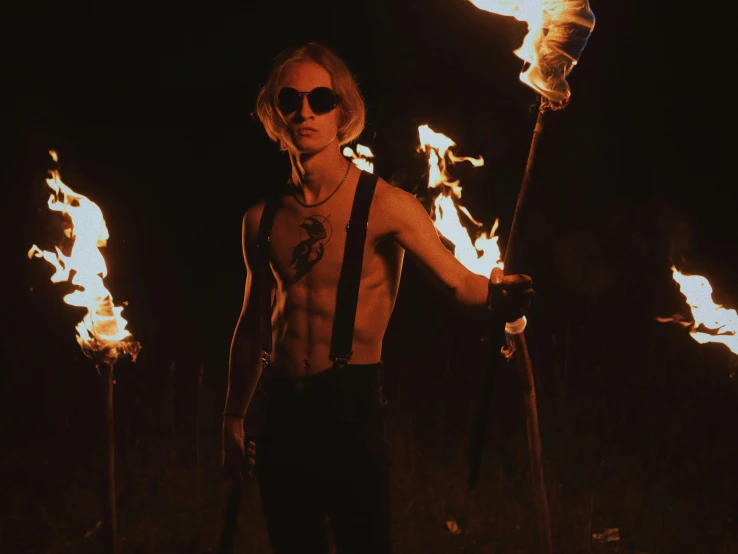 a man standing in the middle of a field holding torches, an album cover, inspired by James Bolivar Manson, trending on pexels, young blonde boy fantasy thief, flames around body, frank dillane, welcome to the circus