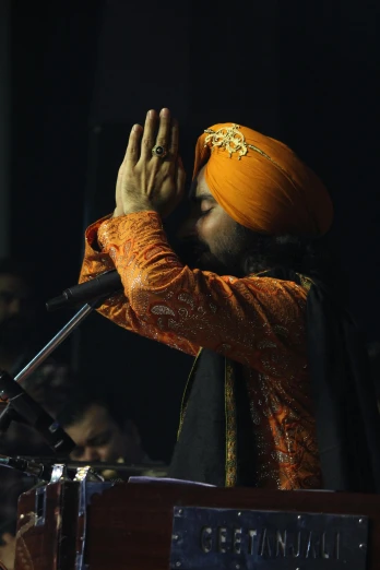 a man in a turban standing in front of a microphone, an album cover, by Manjit Bawa, pexels, praying, an orange, blessing hands, recital