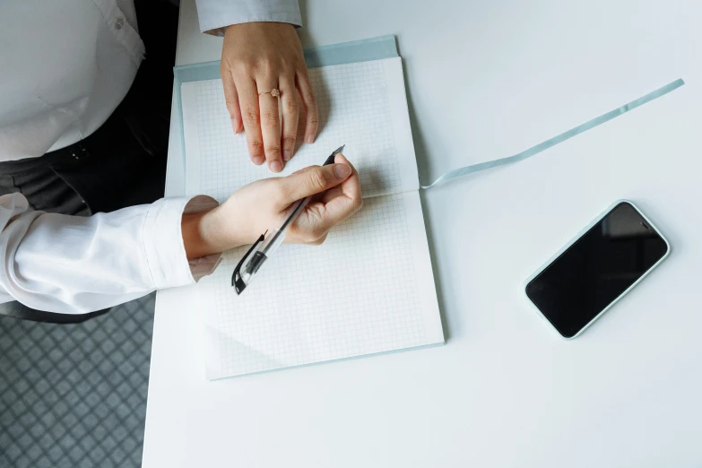 a person sitting at a table writing on a piece of paper, trending on pexels, wearing lab coat and a blouse, taken on iphone 14 pro, minimalist desk, thumbnail