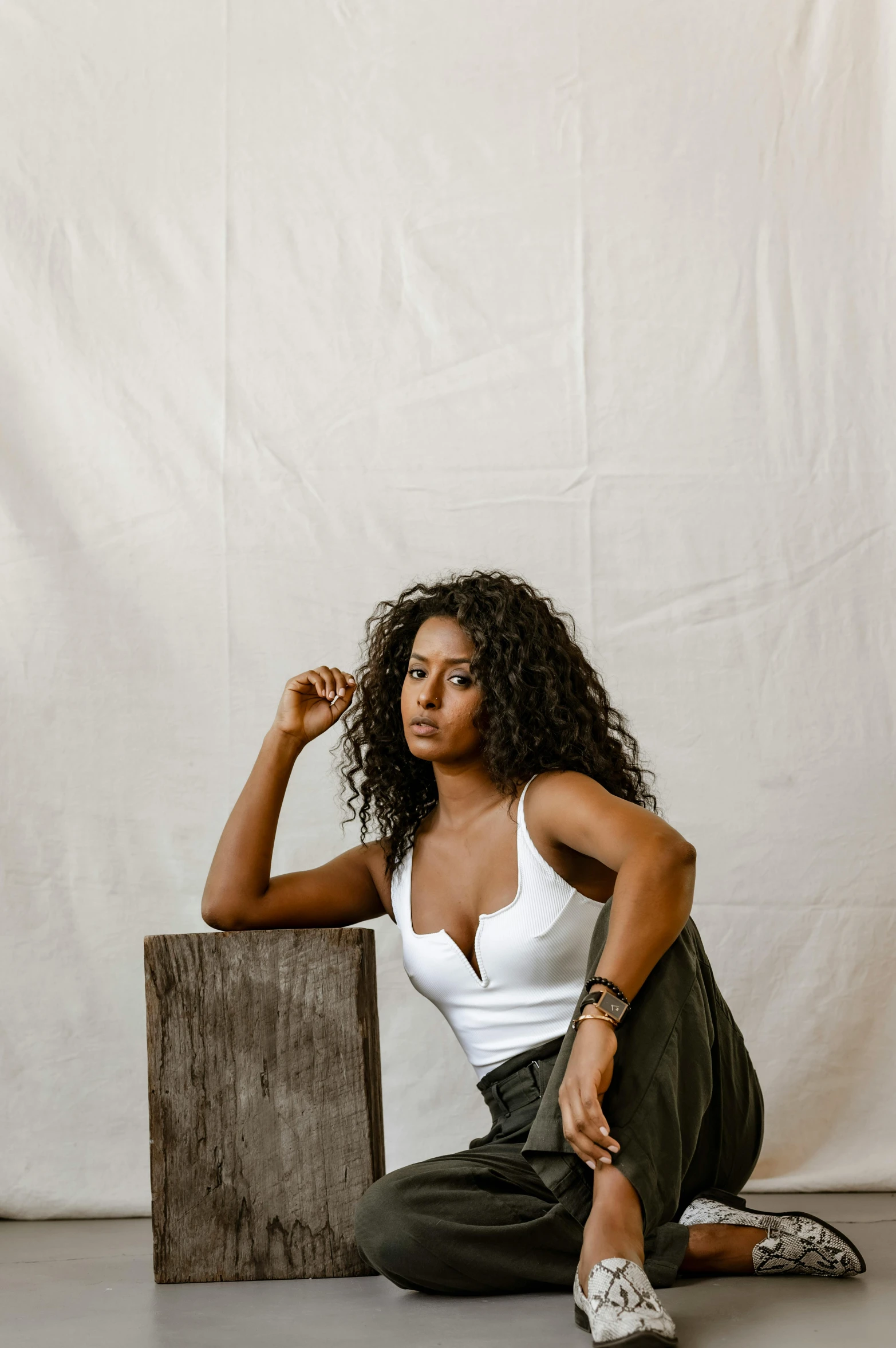 a woman sitting on the floor in front of a white backdrop, inspired by Esaias Boursse, trending on pexels, curls on top, tanned ameera al taweel, seated on wooden chair, from waist up