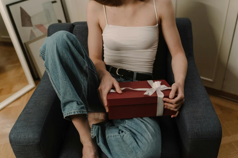 a woman sitting in a chair holding a red box, pexels contest winner, jeans and t shirt, birthday wrapped presents, thigh gap, maroon and white