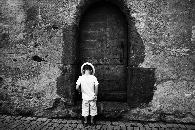 a little boy that is standing in front of a door, by Giuseppe Camuncoli, white hat, back and white, near a stone gate, lowres