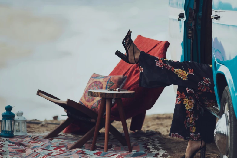 a woman sitting on a blanket in front of a camper, an album cover, trending on unsplash, arabesque, a woman wearing red high heels, seated on wooden chair, near the sea, middle eastern style vendors