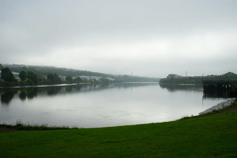a large body of water sitting on top of a lush green field, a picture, under a gray foggy sky, glasgow, lakeside, urban surroundings