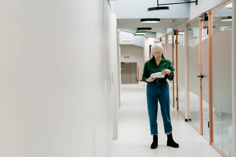 a woman standing in a hallway holding a piece of paper, by Emma Andijewska, pexels contest winner, modernism, in a open-space working space, old man, healthcare, full body length