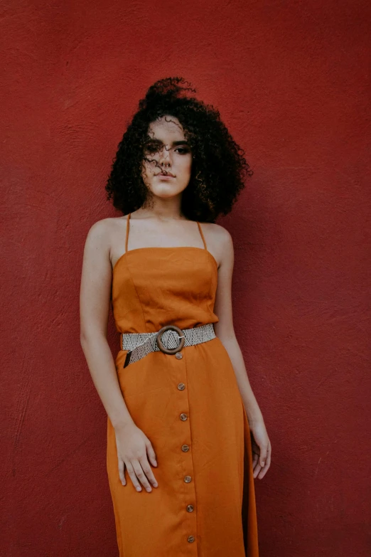 a woman standing in front of a red wall, an album cover, trending on pexels, wearing orange sundress, curly hair, cinematic outfit photo, square