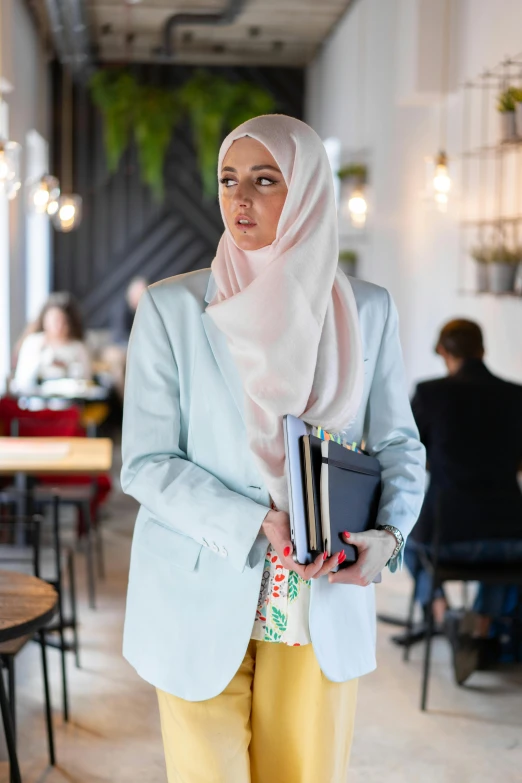 a woman in a hijab is holding a book, trending on pexels, wearing a light blue suit, standing in a restaurant, pastel clothing, wearing jacket and skirt