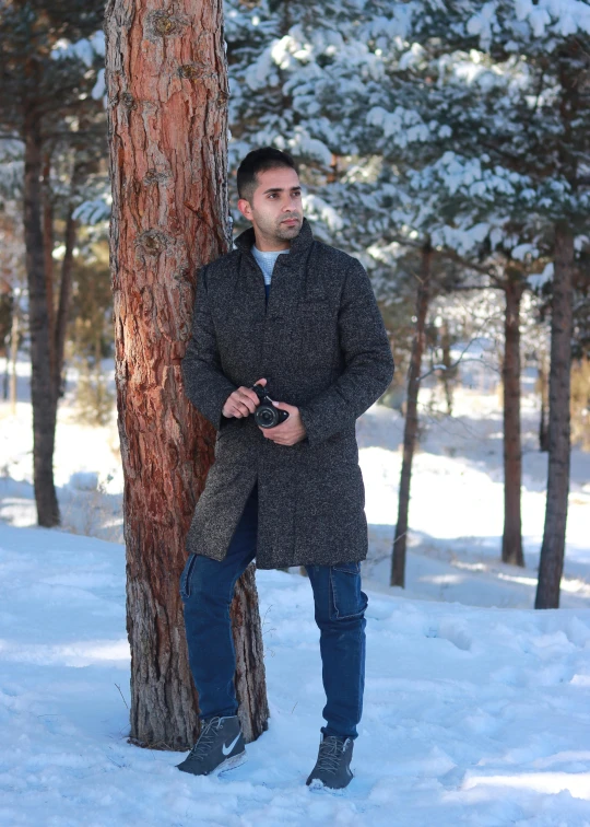 a man standing next to a tree in the snow, full body profile camera shot, in avila pinewood, wearing a long coat, profile image