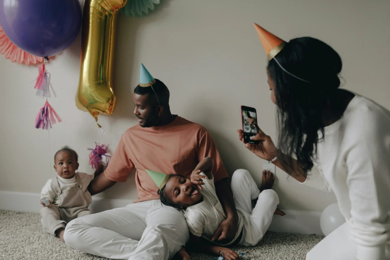 a woman taking a picture of a family sitting on the floor, by Carey Morris, pexels contest winner, hyperrealism, party balloons, jaylen brown, caring fatherly wide forehead, wearing a party hat